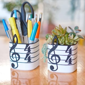 May include: Two white cylindrical containers with musical note and 4/4 time signature designs. One holds pens, pencils, and scissors; the other contains succulents. The containers sit on a wooden surface.