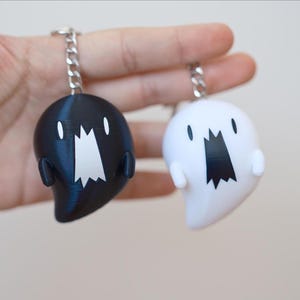 May include: Two ghost-shaped keychains, one black and one white, each with a silver chain. The ghosts have a wide, open mouth design and small, rounded arms. The keychains are held against a plain background.