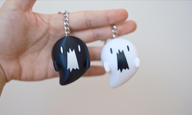 May include: Two ghost-shaped keychains, one black and one white, each with a silver chain. The ghosts have a wide, open mouth design and small, rounded arms. The keychains are held against a plain background.