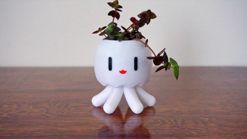 May include: White ceramic planter shaped like an octopus with a happy face and red lips. The planter has a small plant with green and burgundy leaves growing inside.