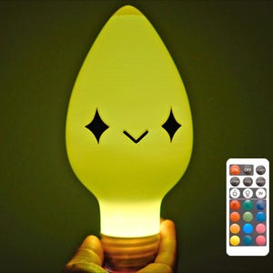 May include: A yellow light bulb-shaped lamp with a smiling face and a remote control. The lamp has a warm glow and the remote has multiple colour options. The lamp is held by a hand.