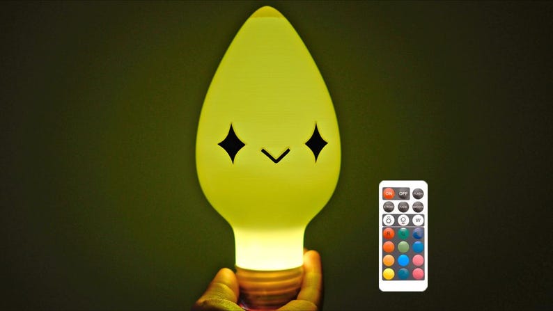 May include: A yellow light bulb-shaped lamp with a smiling face and a remote control. The lamp has a warm glow and the remote has multiple colour options. The lamp is held by a hand.