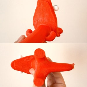 May include: A bright orange, 3D-printed octopus-like sculpture with white spherical eyes. The sculpture has a textured surface and is held in a person's hand. The design features a central body with multiple arms and a unique, artistic aesthetic.