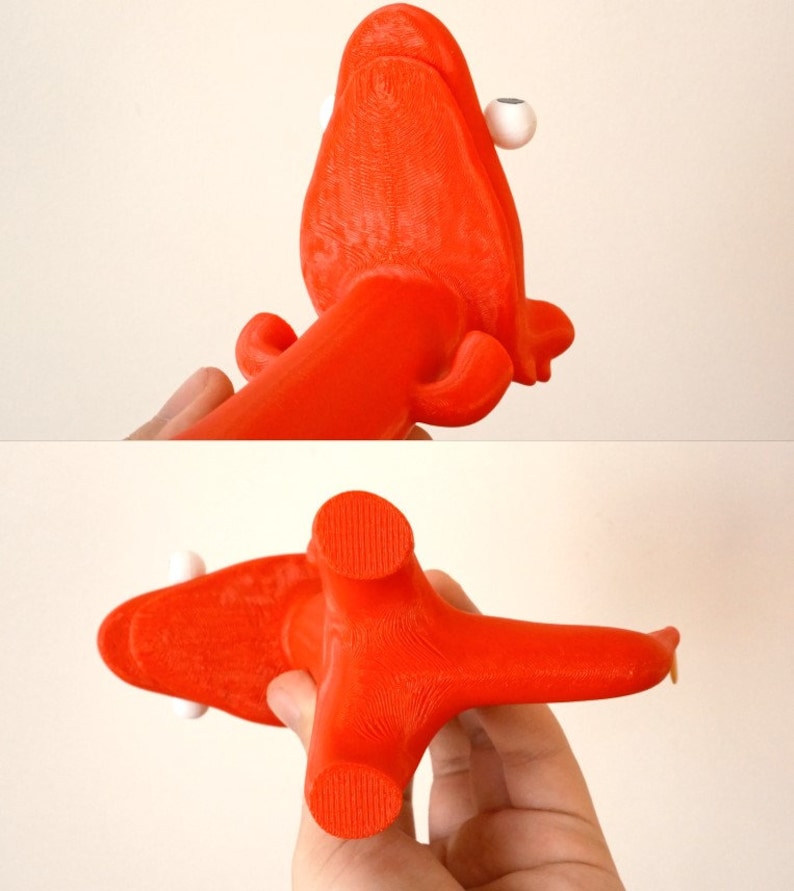 May include: A bright orange, 3D-printed octopus-like sculpture with white spherical eyes. The sculpture has a textured surface and is held in a person's hand. The design features a central body with multiple arms and a unique, artistic aesthetic.