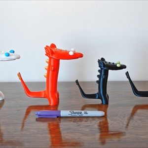 May include: Four whimsical dragon figurines in white, red, and black, each with a unique design. The dragons have stylized features and are arranged on a wooden surface. A purple Sharpie marker is in the foreground.