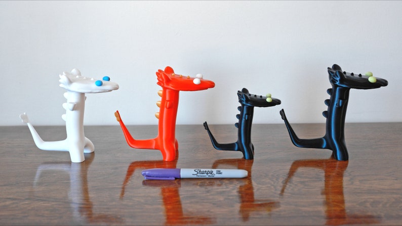 May include: Four whimsical dragon figurines in white, red, and black, each with a unique design. The dragons have stylized features and are arranged on a wooden surface. A purple Sharpie marker is in the foreground.