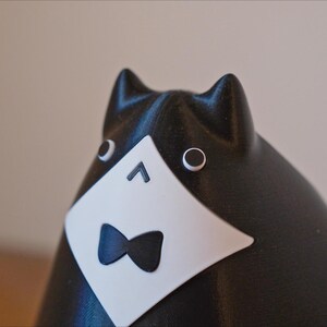 May include: A black and white cat figurine with a tuxedo design. The cat has a white bib with a black bow tie, and round eyes. The ears are pointed, and the figurine has a smooth finish. The cat is a decorative item.