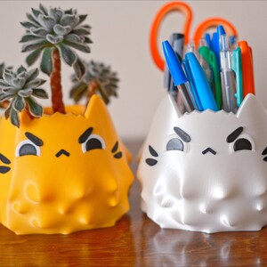 May include: Two 3D printed cat-shaped planters, one yellow with a succulent plant and the other white with a collection of pens and pencils.