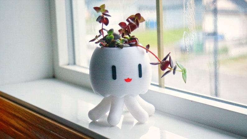 May include: White ceramic planter shaped like an octopus with a happy face and red lips. The planter is holding a small succulent plant with green leaves and red stems.