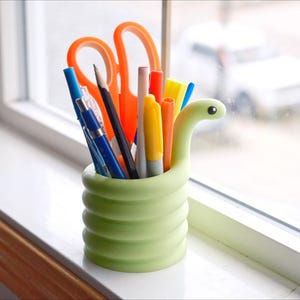 May include: A light green, snake-shaped desk organizer filled with pens, pencils, and orange scissors. The organizer sits on a white windowsill, with a blurred view of a car and the outdoors in the background.