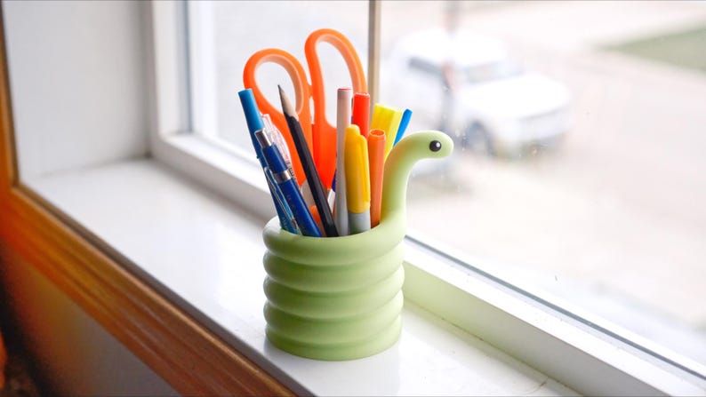 May include: A light green, snake-shaped desk organizer filled with pens, pencils, and orange scissors. The organizer sits on a white windowsill, with a blurred view of a car and the outdoors in the background.