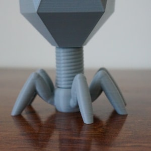 May include: A gray 3D printed model of a bacteriophage, a type of virus that infects bacteria. The model has a hexagonal head, a cylindrical neck, and six legs.