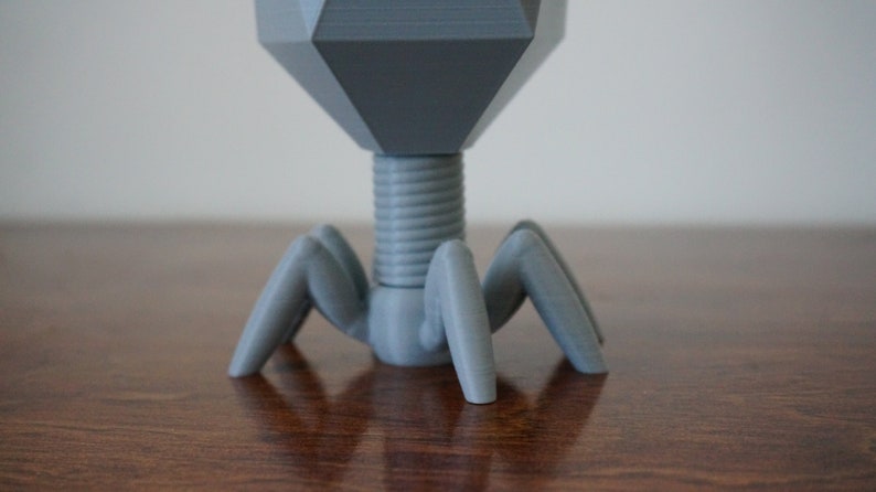 May include: A gray 3D printed model of a bacteriophage, a type of virus that infects bacteria. The model has a hexagonal head, a cylindrical neck, and six legs.