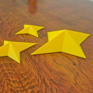 May include: Three yellow paper stars of different sizes on a wooden surface.