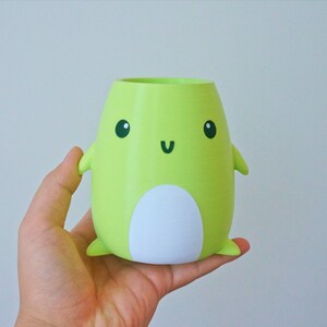 May include: A green ceramic planter shaped like a cartoon character with a white belly and black eyes and mouth. The planter has small arms and legs.