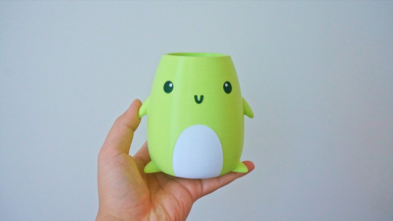 May include: A green ceramic planter shaped like a cartoon character with a white belly and black eyes and mouth. The planter has small arms and legs.
