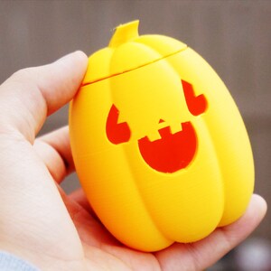 May include: A yellow plastic pumpkin with a red smiling face carved into it. The pumpkin is being held in a person's hand.