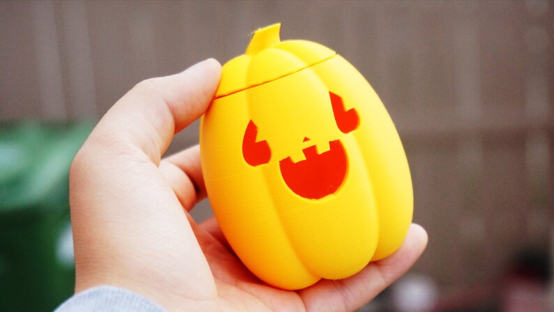 May include: A yellow plastic pumpkin with a red smiling face carved into it. The pumpkin is being held in a person's hand.