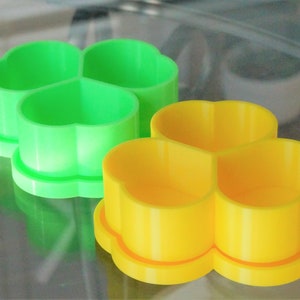 May include: Two plastic clover-shaped cookie cutters. One is bright green, the other is yellow. The cutters have a three-leaf clover design and are sitting on a reflective surface. These are likely for baking or crafting.