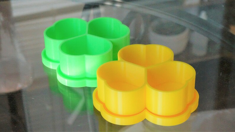 May include: Two plastic clover-shaped cookie cutters. One is bright green, the other is yellow. The cutters have a three-leaf clover design and are sitting on a reflective surface. These are likely for baking or crafting.