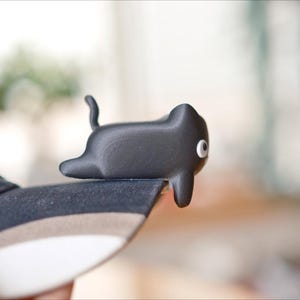 May include: A black cat-shaped shoe accessory with a white eye detail rests on the edge of a navy blue trainer. The accessory has a curved tail and is made of a smooth material. The trainer has the brand name "FILA" on the side.