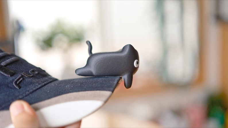 May include: A black cat-shaped shoe accessory with a white eye detail rests on the edge of a navy blue trainer. The accessory has a curved tail and is made of a smooth material. The trainer has the brand name "FILA" on the side.