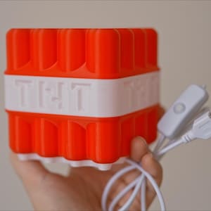 May include: A red and white 3D printed cube with the word "TNT" on the side. The cube is connected to a white USB cable with a switch.