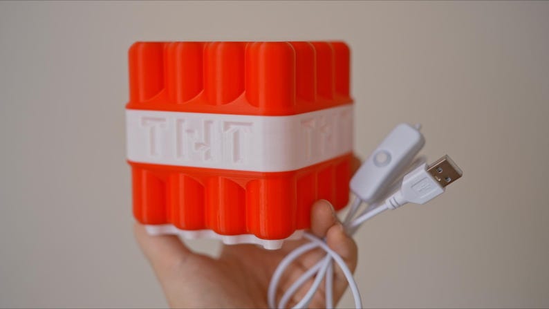 May include: A red and white 3D printed cube with the word "TNT" on the side. The cube is connected to a white USB cable with a switch.