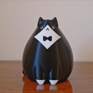 May include: A black cat figurine with a white bib and bow tie. The cat has white feet and ears. The figurine is sitting on a wooden surface.