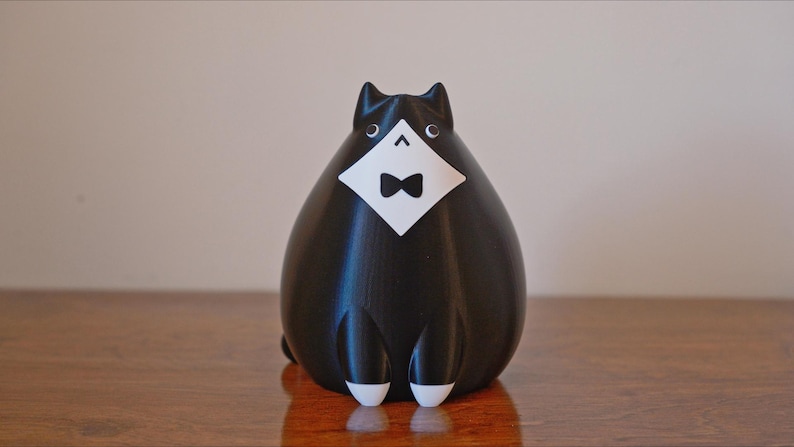 May include: A black cat figurine with a white bib and bow tie. The cat has white feet and ears. The figurine is sitting on a wooden surface.