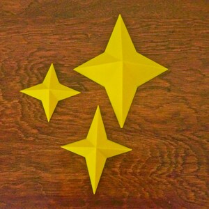 May include: Three yellow paper stars of different sizes on a brown wooden background.