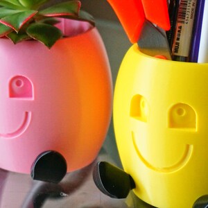 May include: Two colorful, smiling planters with black legs. One planter is pink and holds a small succulent plant. The other planter is yellow and holds pens and scissors.