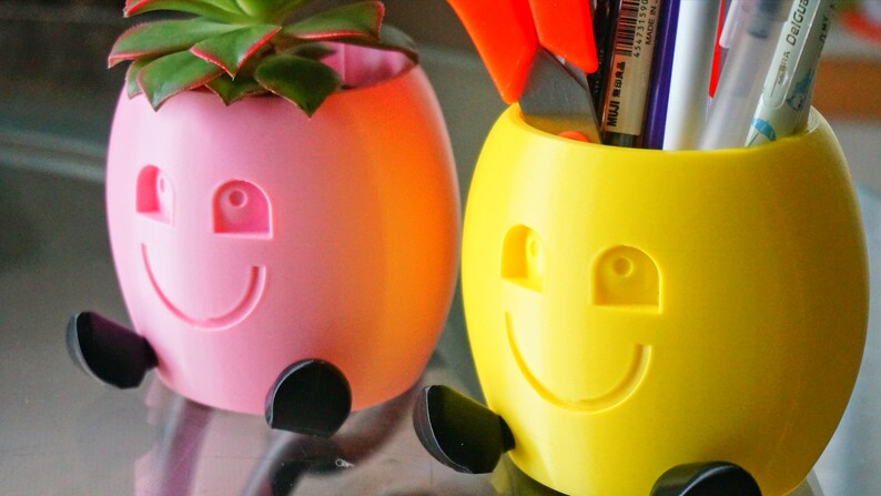 May include: Two colorful, smiling planters with black legs. One planter is pink and holds a small succulent plant. The other planter is yellow and holds pens and scissors.