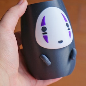 May include: A black tumbler featuring a white face with purple accents, resembling a character from an animated film. The tumbler has small gray appendages and is held in a hand.