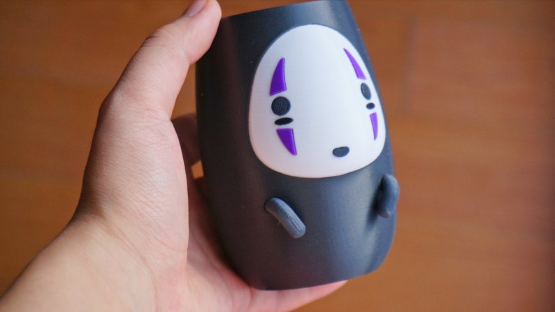 May include: A black tumbler featuring a white face with purple accents, resembling a character from an animated film. The tumbler has small gray appendages and is held in a hand.