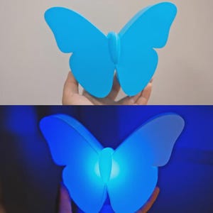 May include: A blue butterfly-shaped lamp. The lamp is held up in front of a neutral background. The lamp is lit up in the second image, with a bright blue glow emanating from the center.