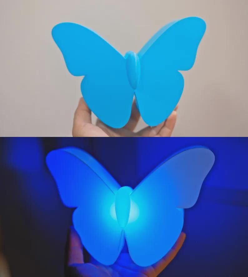 May include: A blue butterfly-shaped lamp. The lamp is held up in front of a neutral background. The lamp is lit up in the second image, with a bright blue glow emanating from the center.