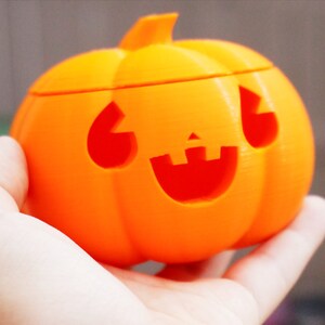 May include: An orange plastic pumpkin with a carved jack-o'-lantern face. The pumpkin is being held in a person's hand.
