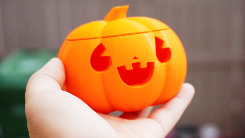 May include: An orange plastic pumpkin with a carved jack-o'-lantern face. The pumpkin is being held in a person's hand.
