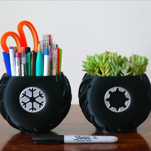 May include: Two black tire-shaped planters, one with a snowflake design and the other with a star design. The planter with the snowflake design is filled with pens, pencils, and scissors. The planter with the star design is filled with a green succulent plant.