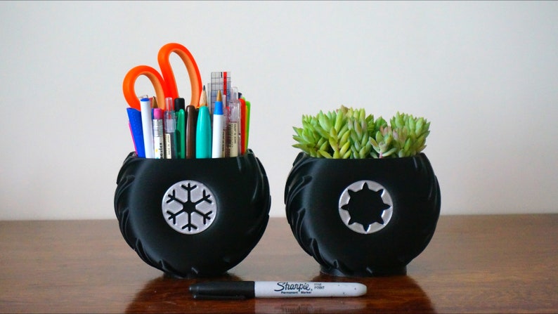 May include: Two black tire-shaped planters, one with a snowflake design and the other with a star design. The planter with the snowflake design is filled with pens, pencils, and scissors. The planter with the star design is filled with a green succulent plant.