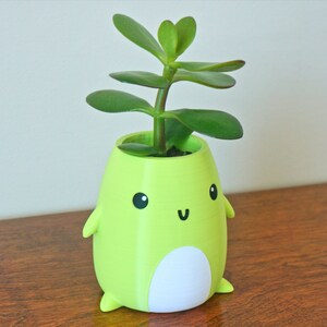 May include: A green ceramic planter shaped like a cartoon character with a white belly and a small green plant growing out of the top.