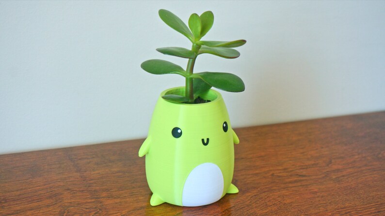 May include: A green ceramic planter shaped like a cartoon character with a white belly and a small green plant growing out of the top.