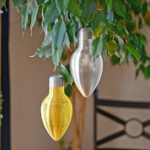 May include: Two decorative light bulb ornaments suspended from a leafy green plant. One bulb is silver, the other is yellow. The bulbs have a metallic finish and are shaped like traditional light bulbs.