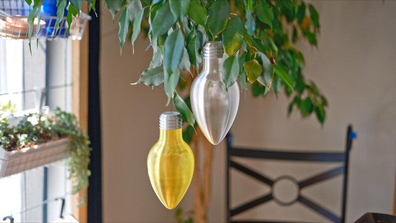 May include: Two decorative light bulb ornaments suspended from a leafy green plant. One bulb is silver, the other is yellow. The bulbs have a metallic finish and are shaped like traditional light bulbs.