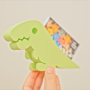 May include: A light green dinosaur-shaped card holder with a cute face. The holder is made of plastic and has a slot for holding cards.