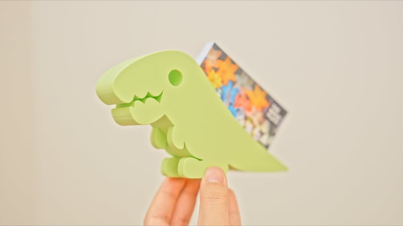 May include: A light green dinosaur-shaped card holder with a cute face. The holder is made of plastic and has a slot for holding cards.