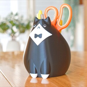 May include: A black and white cat-shaped desk organizer with a bow tie and a white bib. Orange scissors are placed in the back of the organizer. The cat organizer is sitting on a wooden table.