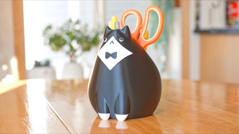May include: A black and white cat-shaped desk organizer with a bow tie and a white bib. Orange scissors are placed in the back of the organizer. The cat organizer is sitting on a wooden table.