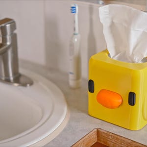 Pu&ograve; includere: Scatola di fazzoletti di plastica gialla a forma di pulcino dei cartoni animati con un becco arancione e occhi neri. La scatola &egrave; piena di fazzoletti bianchi.